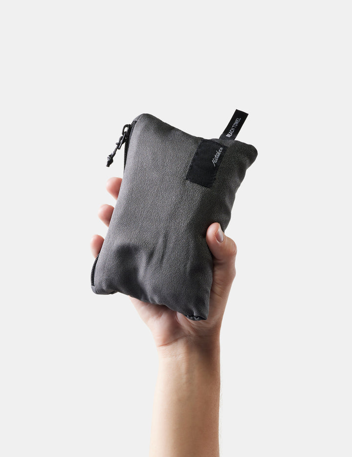 Matador NanoDry™ Packable Beach Towel - hand holding a gray fabric pouch with a black zipper on a white background
