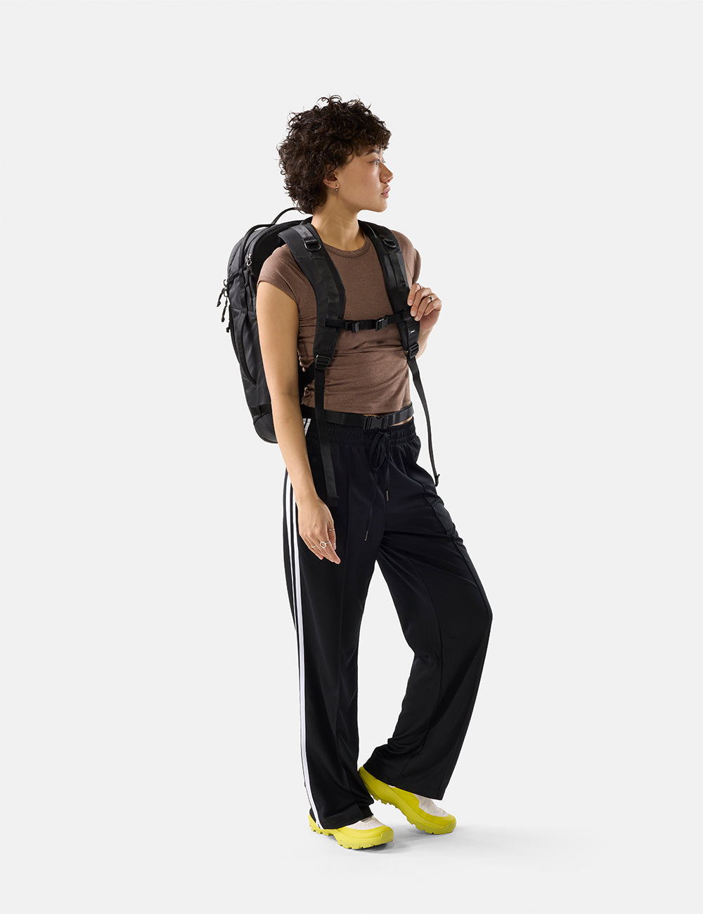 Matador 30L Carry-On Travel Pack in Black - person wearing a brown shirt, black pants with white stripes, and yellow shoes, holding a black backpack on a white background