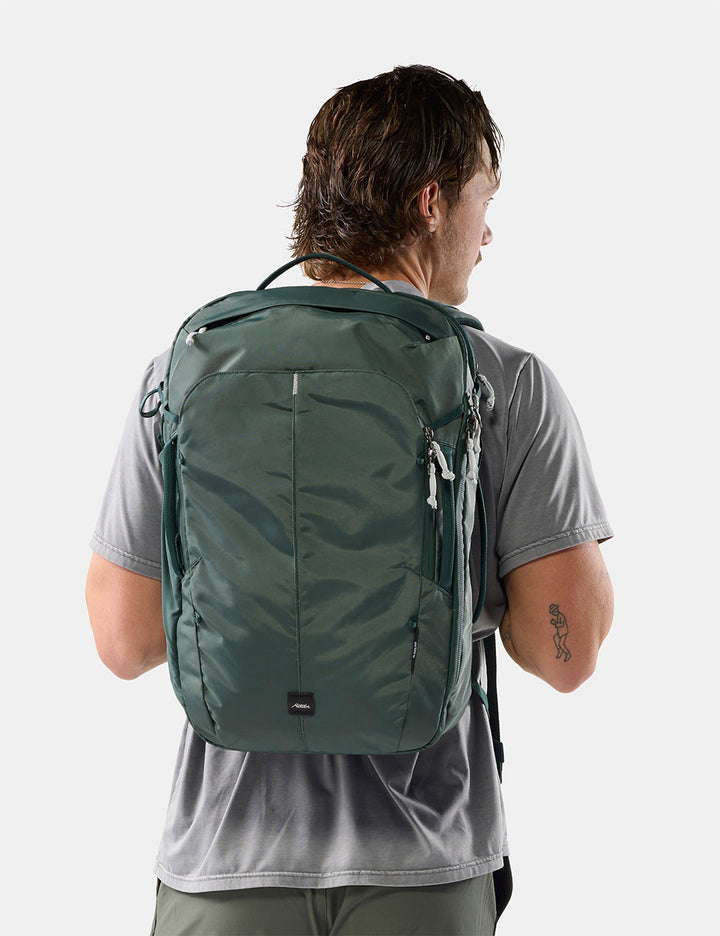Matador 30L Carry-On Travel Pack in Juniper - a man viewed from behind wearing a dark green backpack on his back, paired with a gray t-shirt, against a light gray studio background