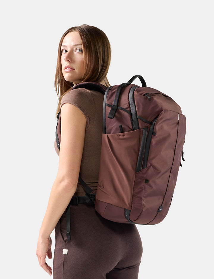 Matador 30L Carry-On Travel Pack in Garnet - a woman wearing a brown sleeveless top and matching leggings is shown from the back wearing a large garnet backpack with black straps and zipper details against a light gray studio background