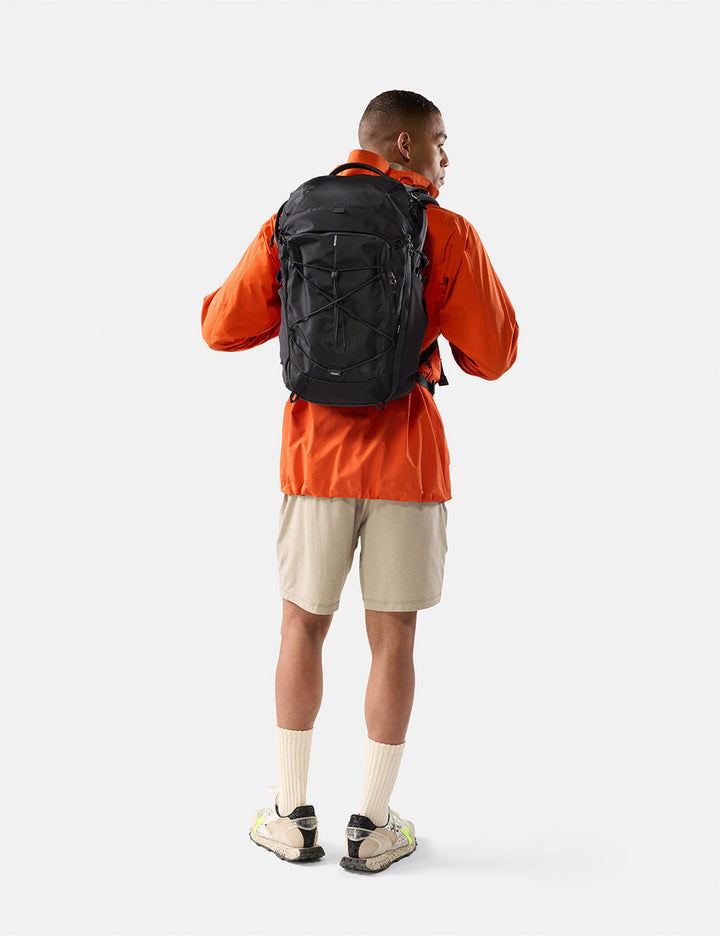 Matador 24L Adventure Pack in Black - person wearing an orange jacket and beige shorts with a black backpack on a white background