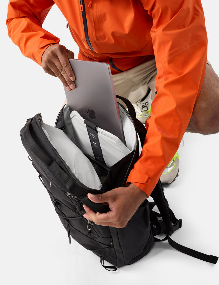 Matador 24L Adventure Pack in Black - person in orange jacket placing a laptop into a black backpack on a white background