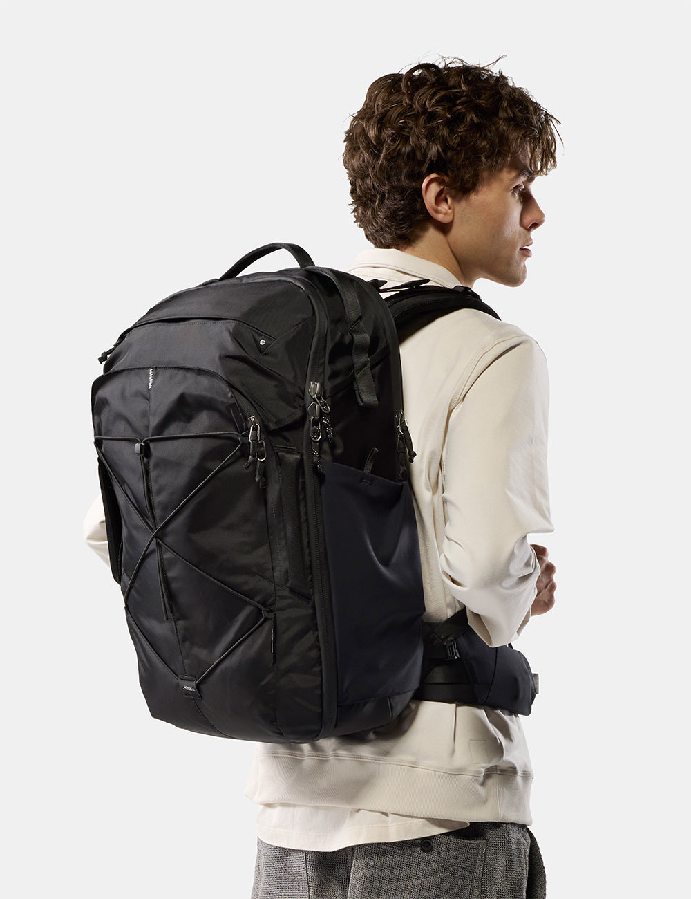 Matador 35L Global Travel Pack in Black - a man wearing a cream-colored hoodie and gray pants is shown from the back and side, carrying a large black technical backpack against a light gray studio background