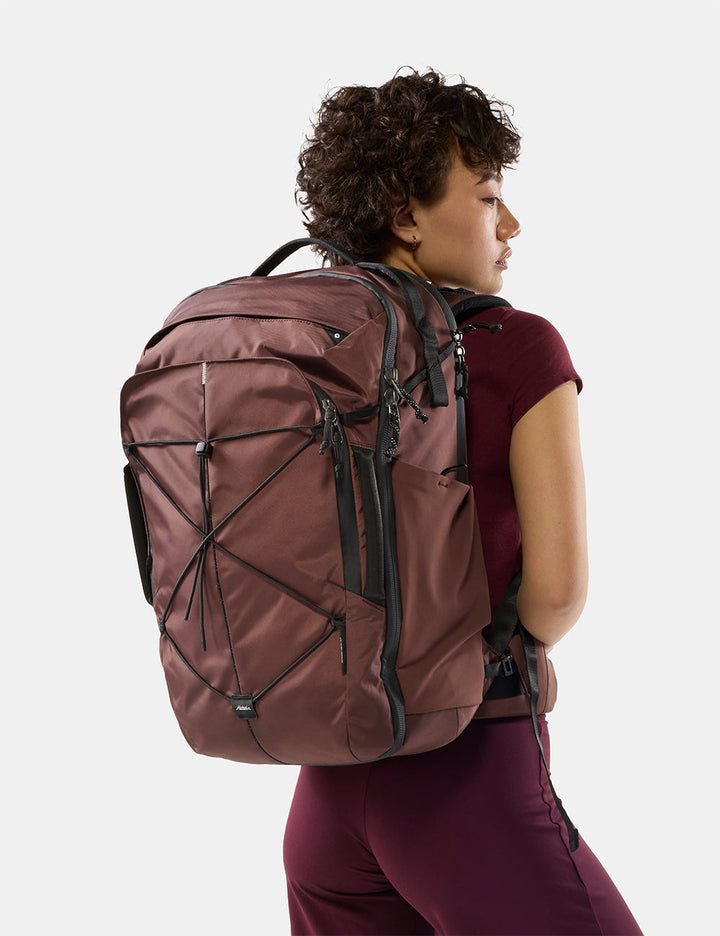 Matador 35L Global Travel Pack in Garnet - a woman wearing a large garnet hiking backpack, shown from the back and side against a clean light gray studio background