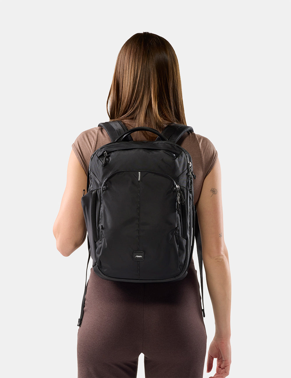 Matador 18L Personal Item Travel Pack in Black - a woman viewed from behind wearing a black backpack against a light gray studio background