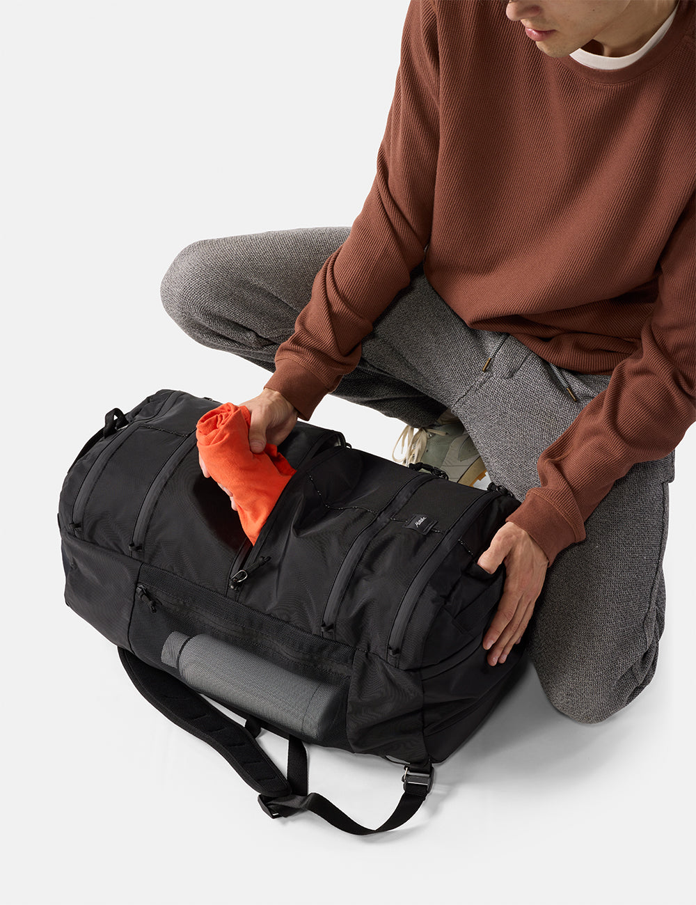 Matador 45L SEG Hybrid Travel Pack in Black - person packing a black duffel bag with an orange item on a light gray background