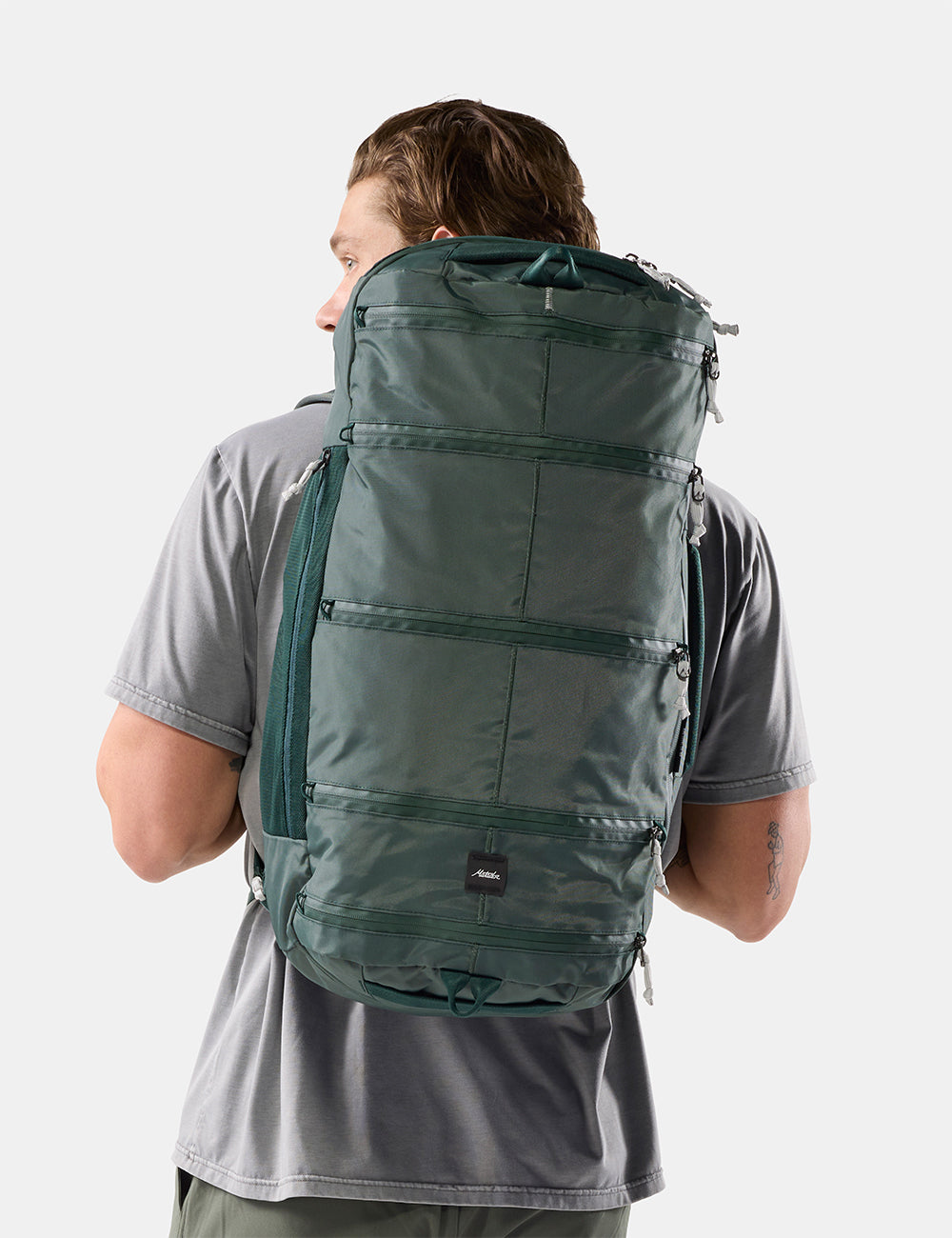 Matador 45L SEG Hybrid Travel Pack in Juniper - a man wearing a gray t-shirt is shown from behind, carrying a large, dark green technical backpack against a light gray studio background