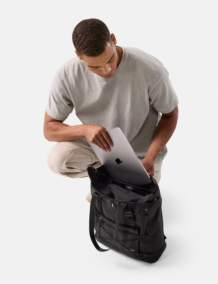 Matador 24L Tote Pack in Black - man placing a laptop into a black tote pack on a white background