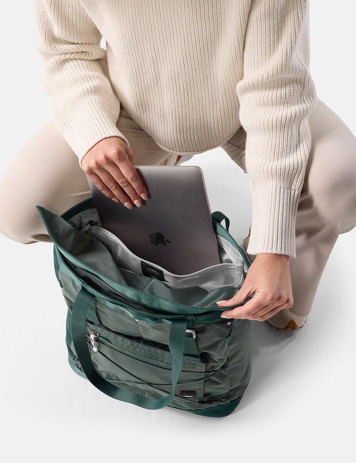 Matador 24L Tote Pack in Juniper - person sitting with laptop and a green tote pack on a white background